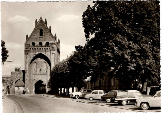 Historisches Tor mit Gebäuden und klassischen Autos in einer ruhigen Straße.