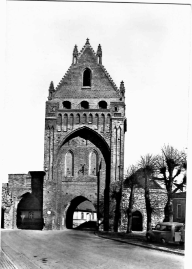 Straße mit historischem Tor, Bäume, alte Gebäude und Busse aus der Mitte des 20. Jahrhunderts.