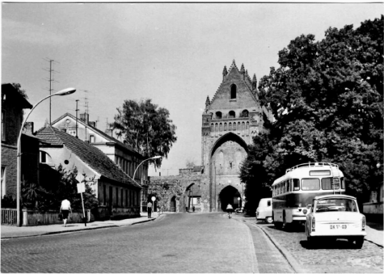 Straße mit historischem Tor, Bäume, alte Gebäude und Busse aus der Mitte des 20. Jahrhunderts.